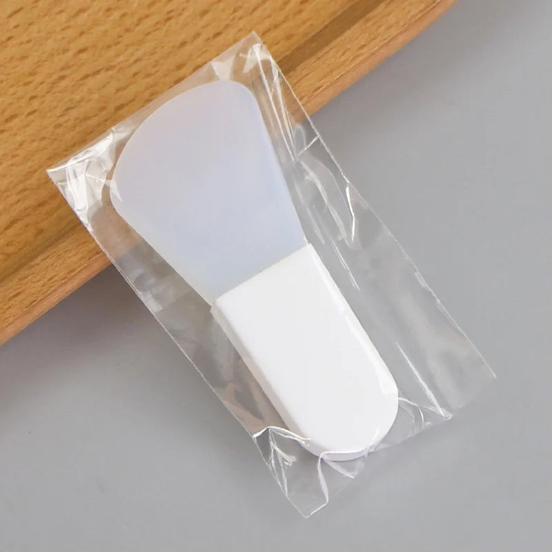 A translucent, flexible silicone facial mask applicator with a white handle, sealed in a clear plastic wrapper.