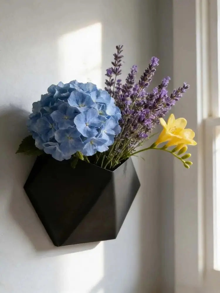 Black hexagonal wall planter holding blue and purple flowers, open top wall-mount design.