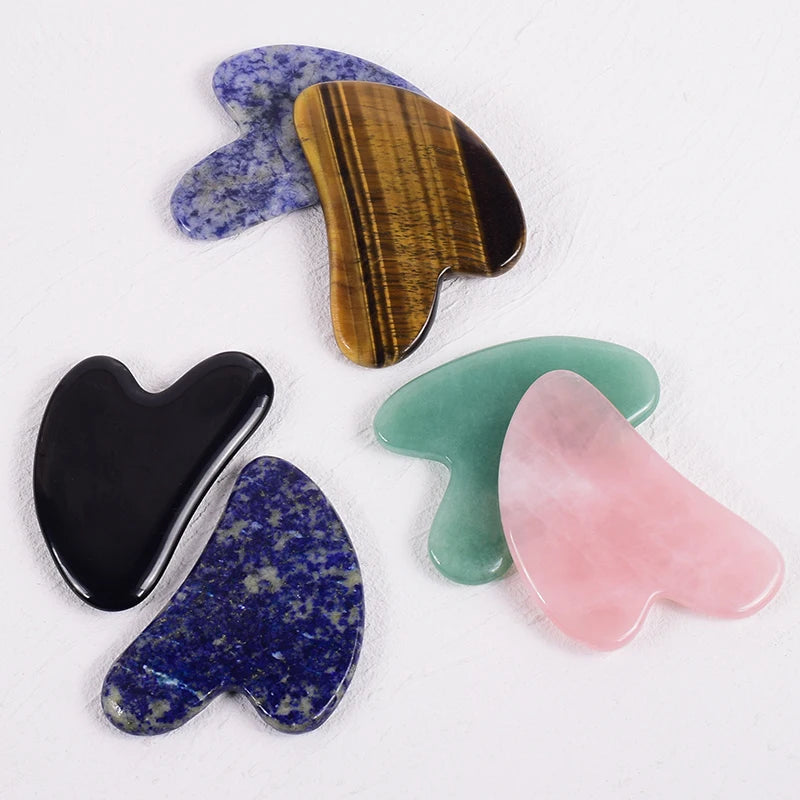 Five heart-shaped gua sha massage tools in various polished gemstones: rose quartz, tiger's eye, black stone, and blue.