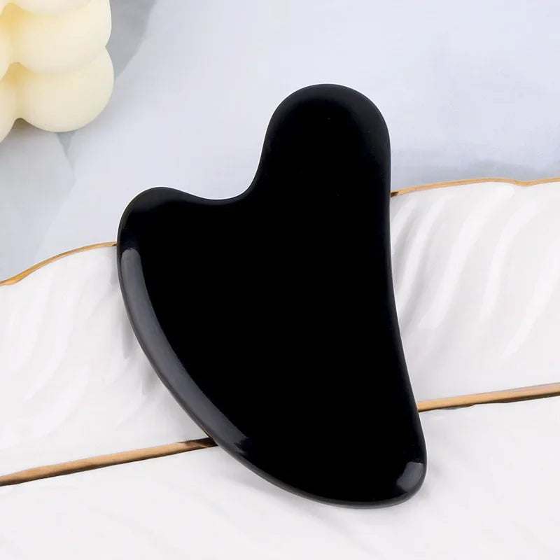 Glossy, heart-shaped black resin gua sha facial massage tool resting on a white, gold-rimmed decorative tray.