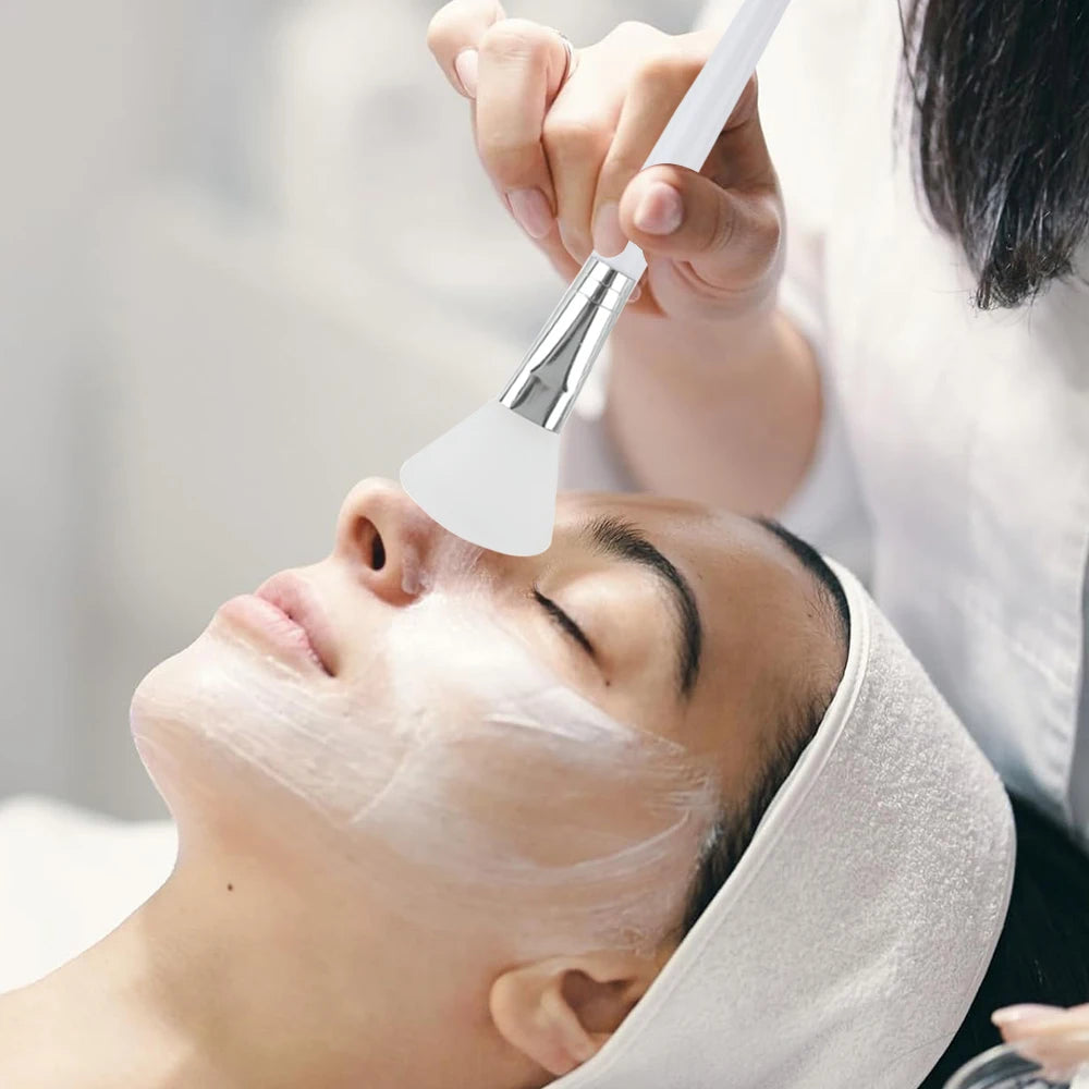 A hand uses a white, flexible silicone brush to apply a facial mask onto a person's skin.