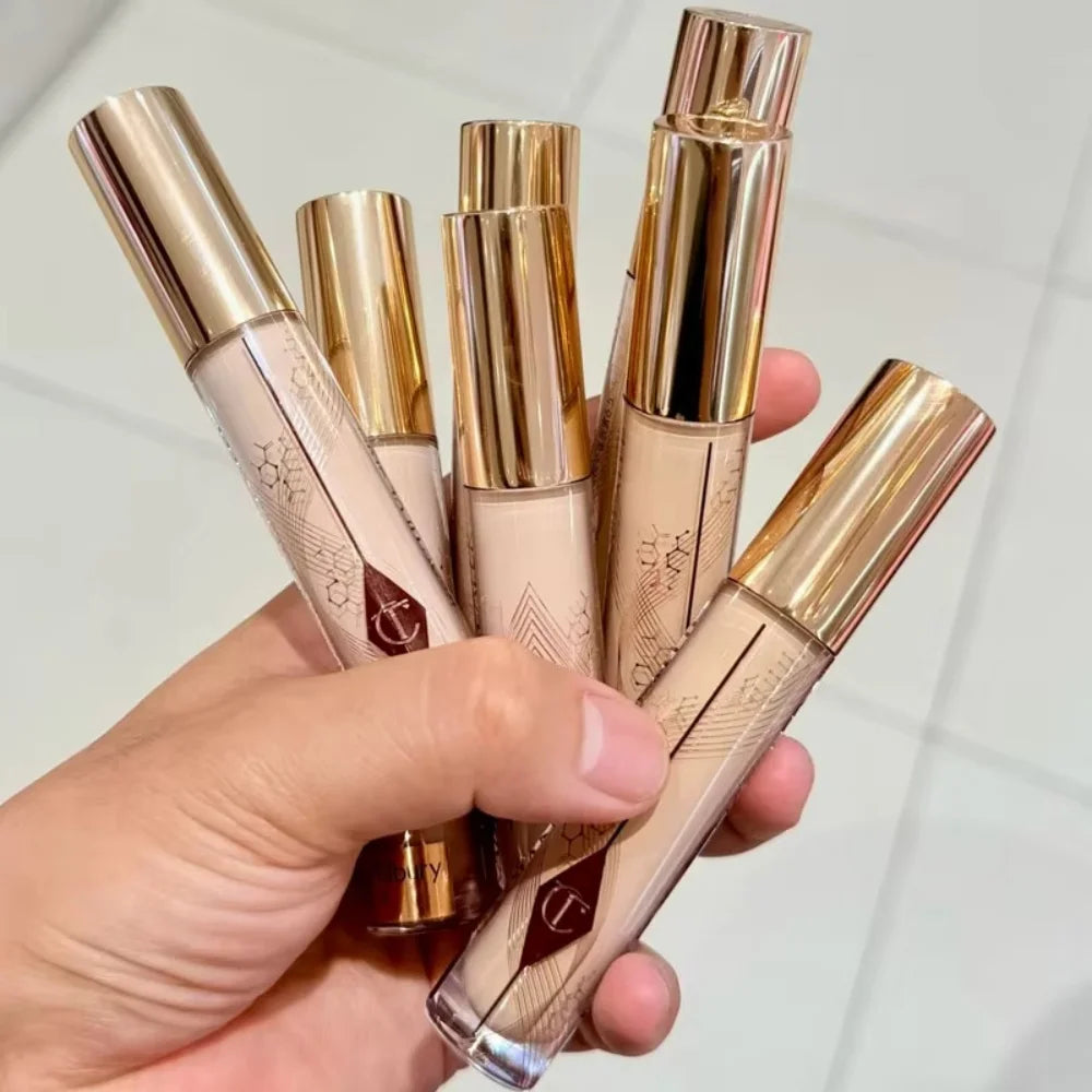 Charlotte Tilbury liquid makeup pens in gold tubes held in hand, featuring a creamy, beige foundation and concealer formula.