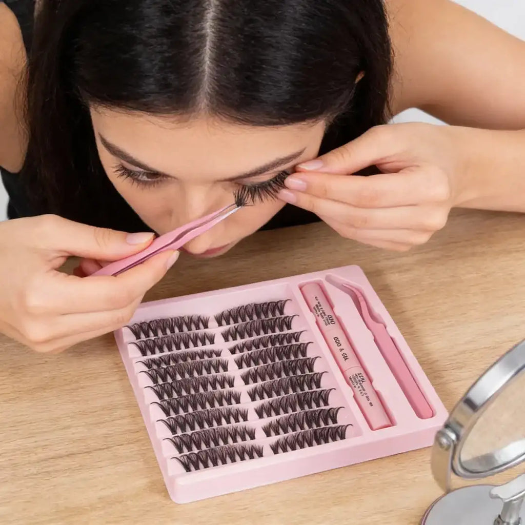 Woman applying natural black DIY lash clusters with pink tweezers from a kit containing bond, seal, and lash tray.