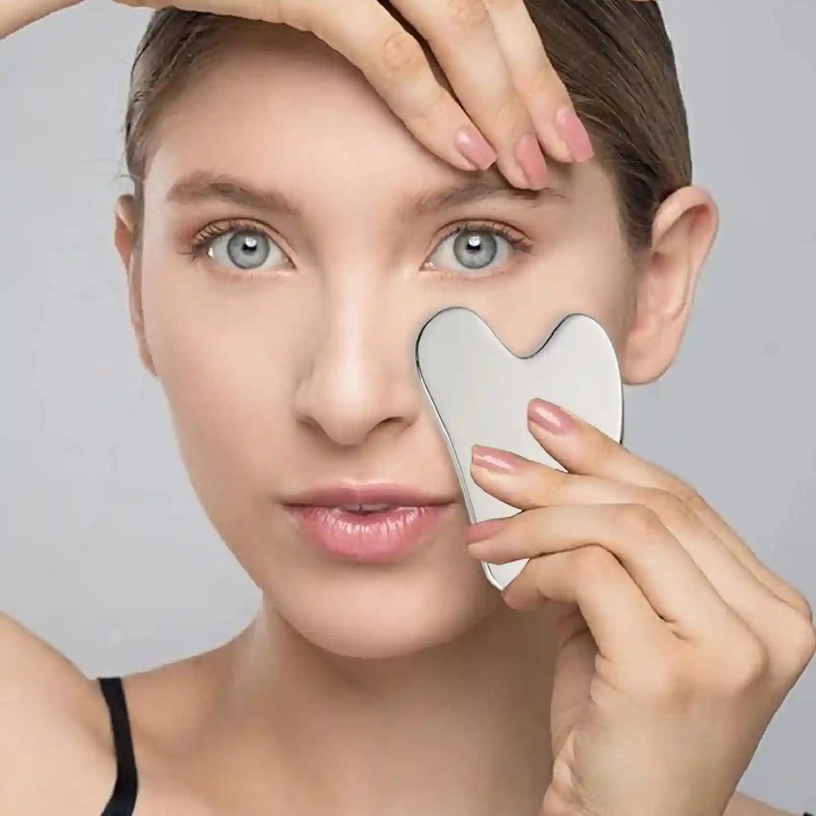 A woman uses a heart-shaped, smooth silver stainless steel Gua Sha tool to massage her cheek.