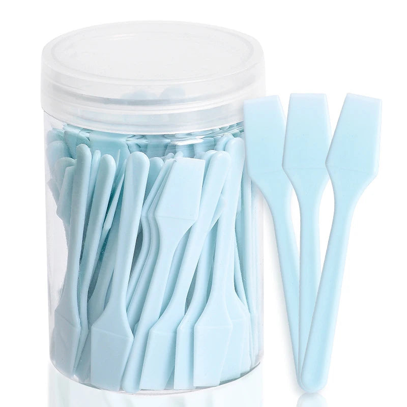 A clear plastic jar filled with 100 mini, light blue plastic cosmetic spatulas for applying creams and masks.