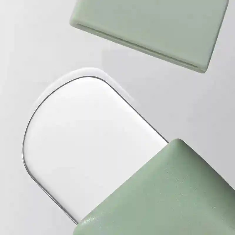 Close-up of a sage green, rounded blackhead removal tool with a thin, curved metal scraper blade on a white background.