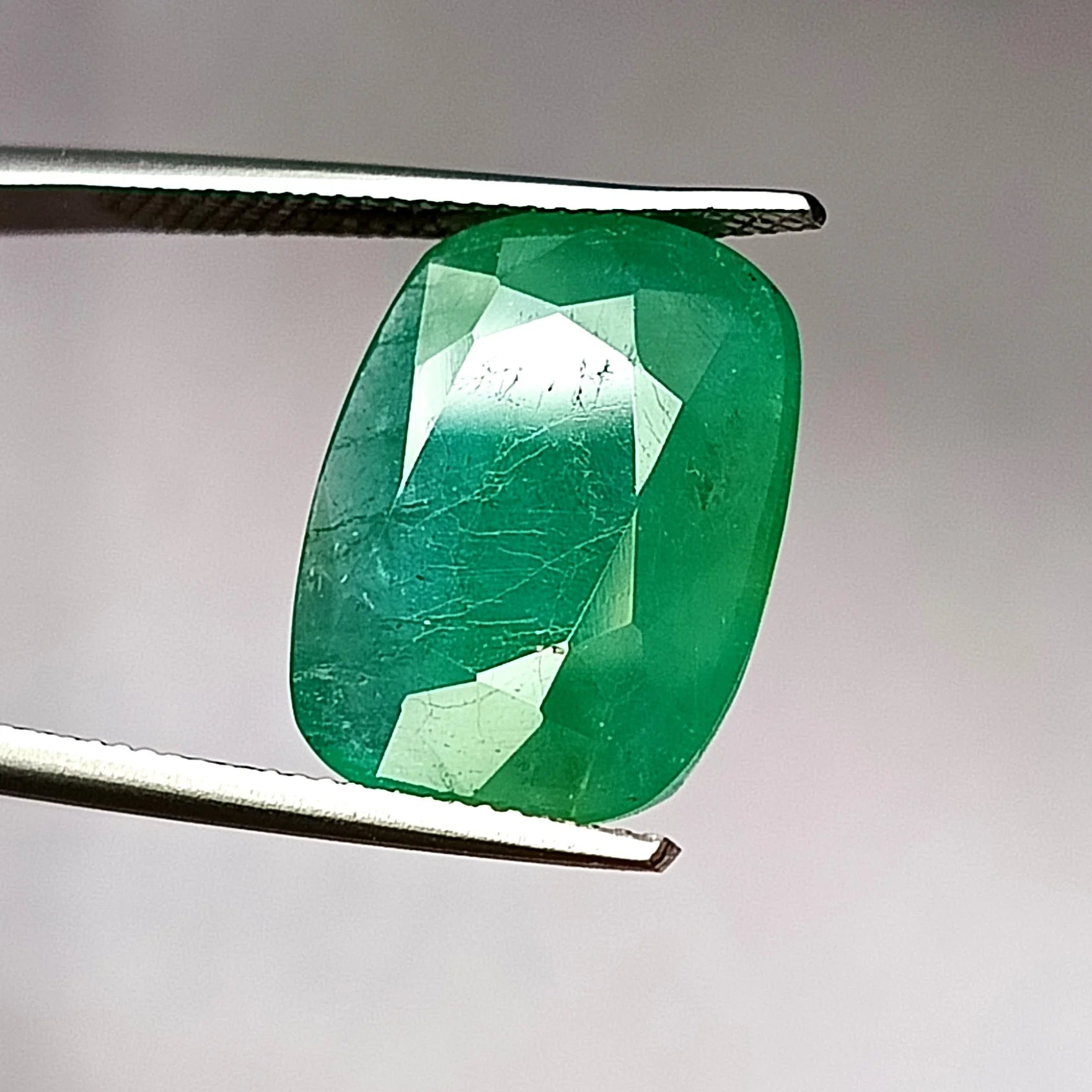 Green oval emerald gemstone, faceted table and facets, set in open tweezers.