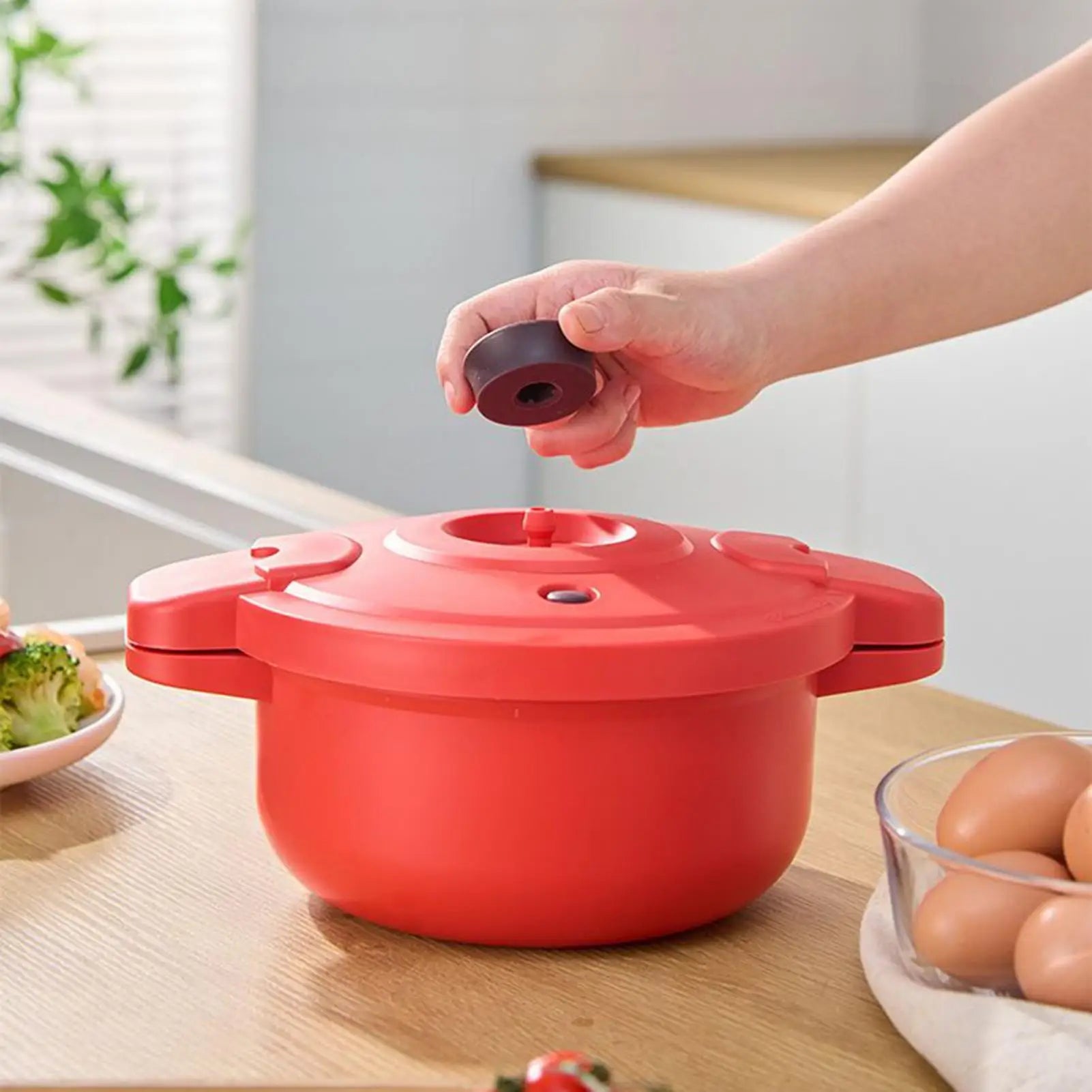 Red microwave pressure cooker pot on table with lid being lifted by hand.