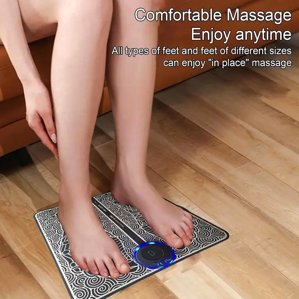 Black and white EMS foot massage mat with blue LED controller, used on wooden floor for sore feet relief.