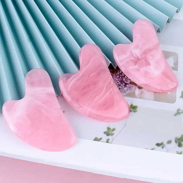 Three heart-shaped, pink marbled resin gua sha massage tools displayed on a light blue and white background.