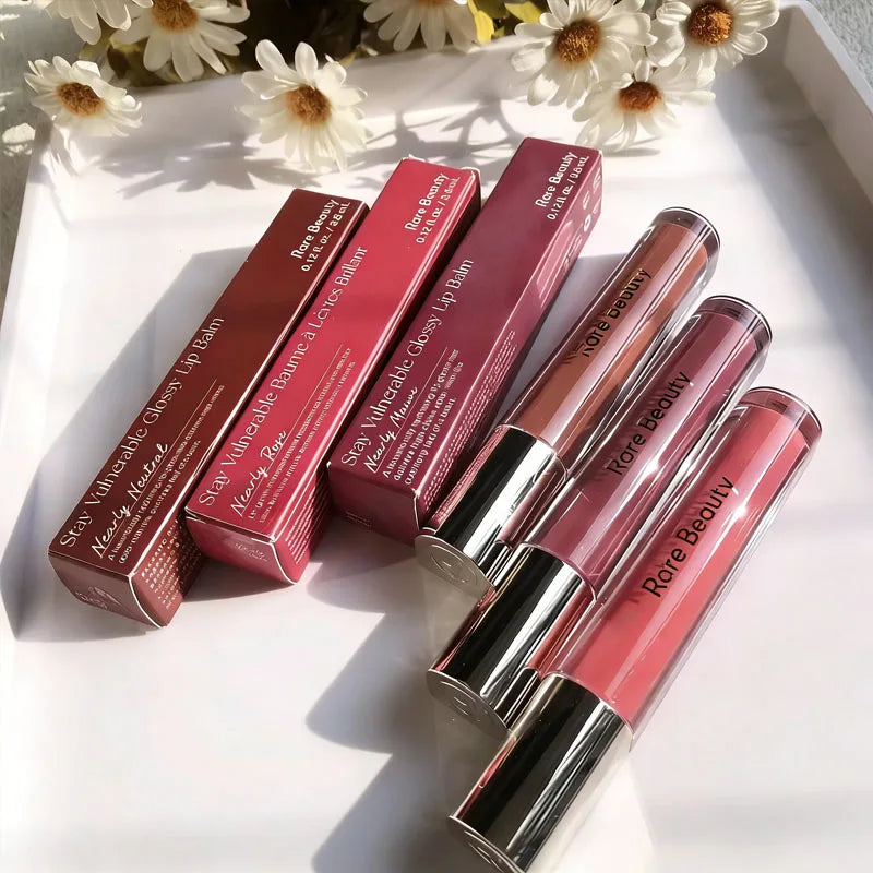Rare Beauty Stay Vulnerable Glossy Lip Balm collection in neutral, rose, and mauve shades with packaging on a white tray.