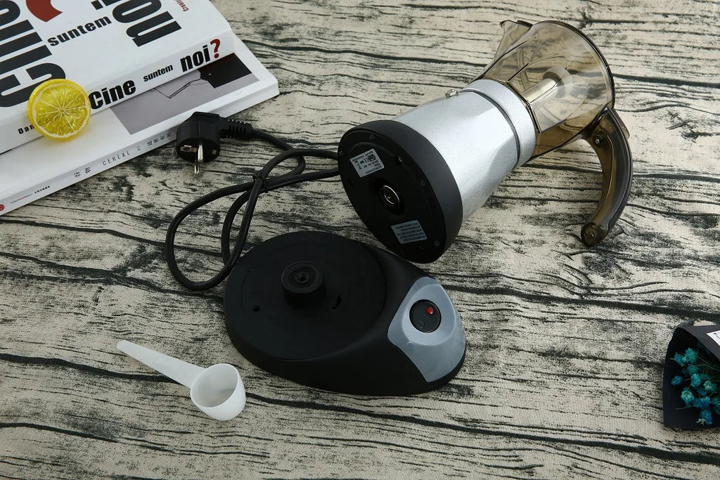 Silver electric moka pot coffee maker with black power base, clear top chamber, and included white plastic scoop.