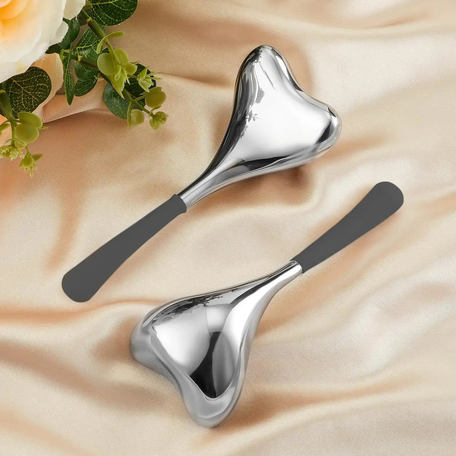 Two heart-shaped stainless steel cryo facial massage sticks with gray handles on a tan satin fabric background.