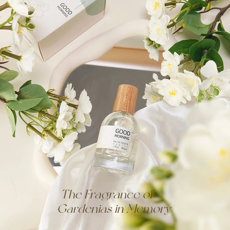 Clear glass perfume bottle with wooden cap on white silk, surrounded by fresh white gardenia flowers.