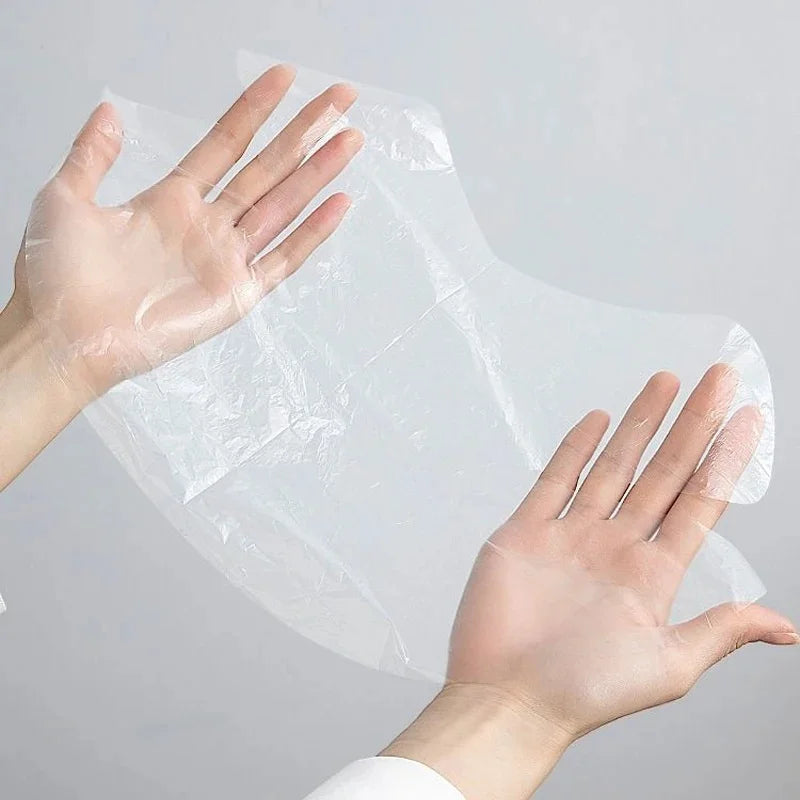 Hands holding a thin, transparent, rectangular plastic face mask film sheet against a plain light gray background.