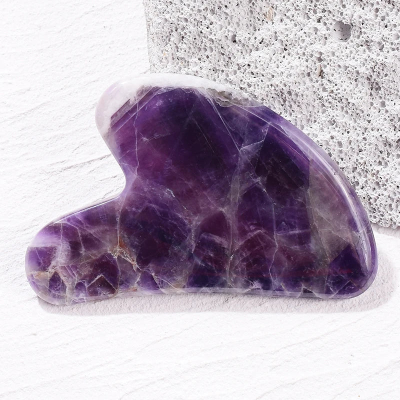 Heart-shaped purple amethyst gua sha tool resting on a white stone surface.