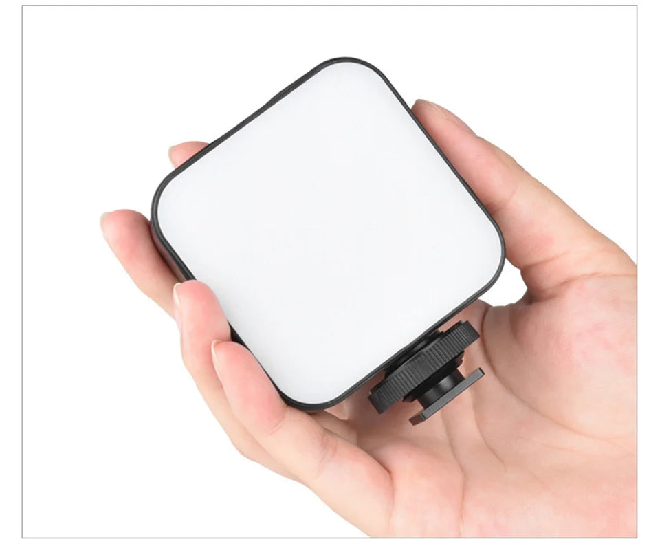 Small, square LED video light with a black frame and cold shoe mount, held in a hand for portable photography lighting.