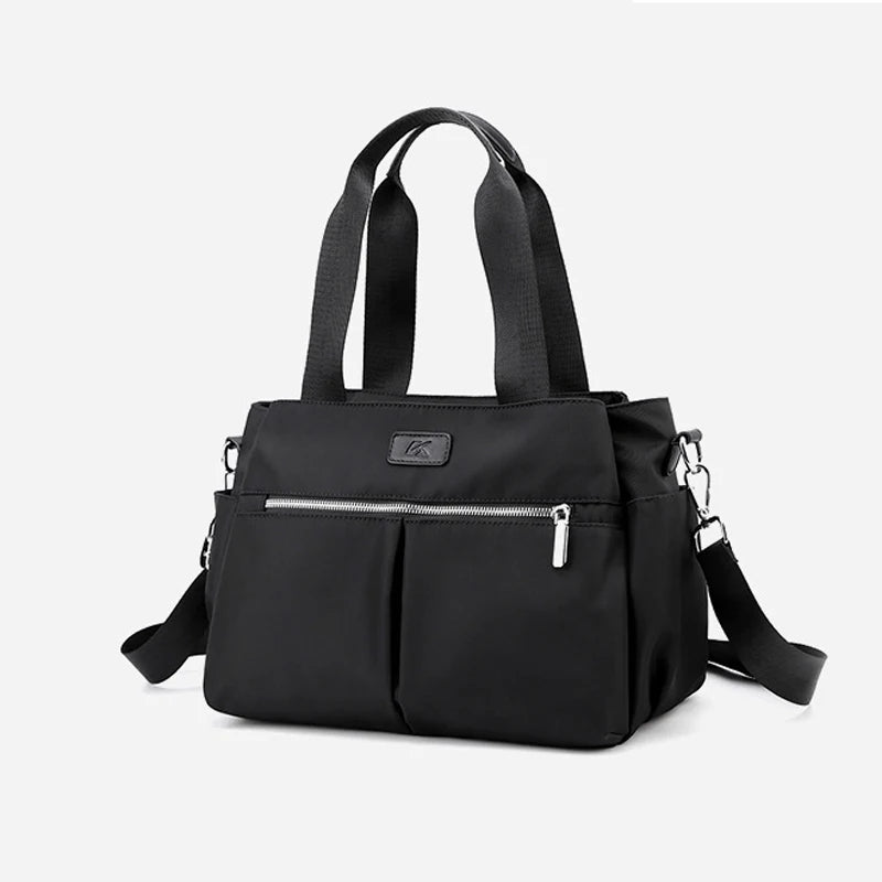 Minimalist black canvas designer handbag with top handles, silver zip pocket, and detachable crossbody shoulder strap.