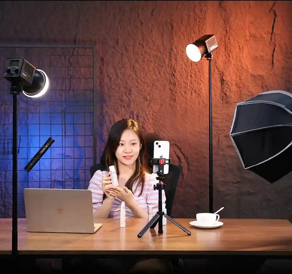 A woman records content on a smartphone mounted on a black tripod, illuminated by studio lights in a home setup.