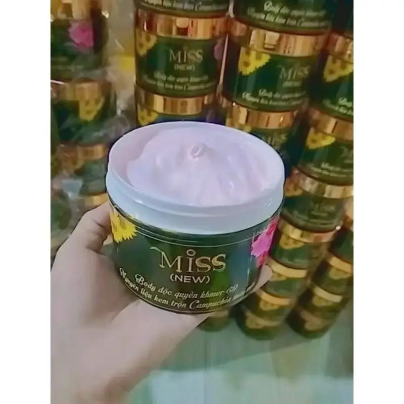 Miss body cream in a dark green jar with a gold lid, filled with thick, light pink moisturizing lotion.