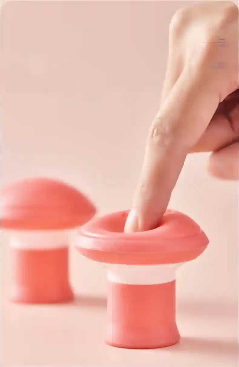 A finger presses into a mushroom-shaped pink and white silicone facial exerciser on a matching pink background.