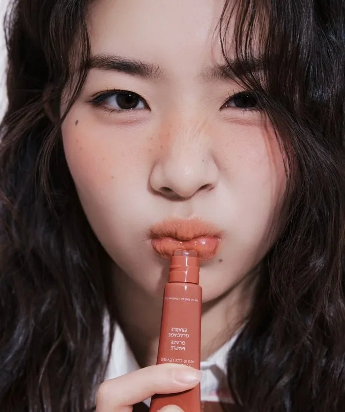 Model applying shiny, maple-toned moisturizing lip glaze from a squeeze tube for a plump, glossy finish.