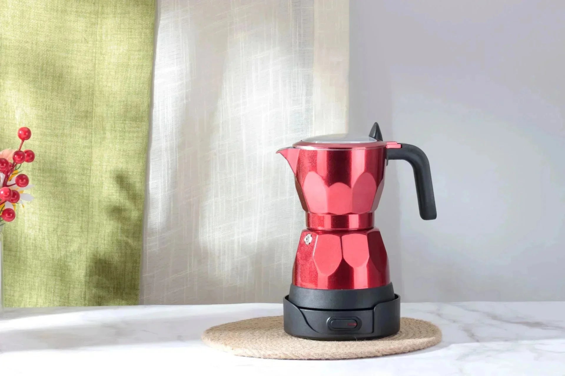 Red metallic electric moka pot coffee maker with black handle and base, standing on a woven placemat.