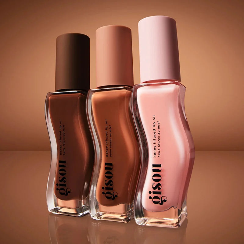 Three Gisou honey-infused lip oil gloss bottles in plum, nude, and pink with a hydrating, plumping, glossy texture.