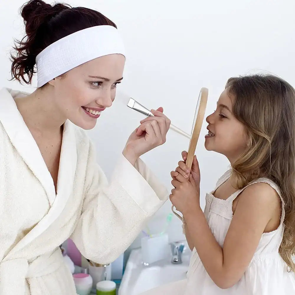 A woman in a robe uses a flat, transparent-handled facial mask brush on her daughter, who holds a small mirror.