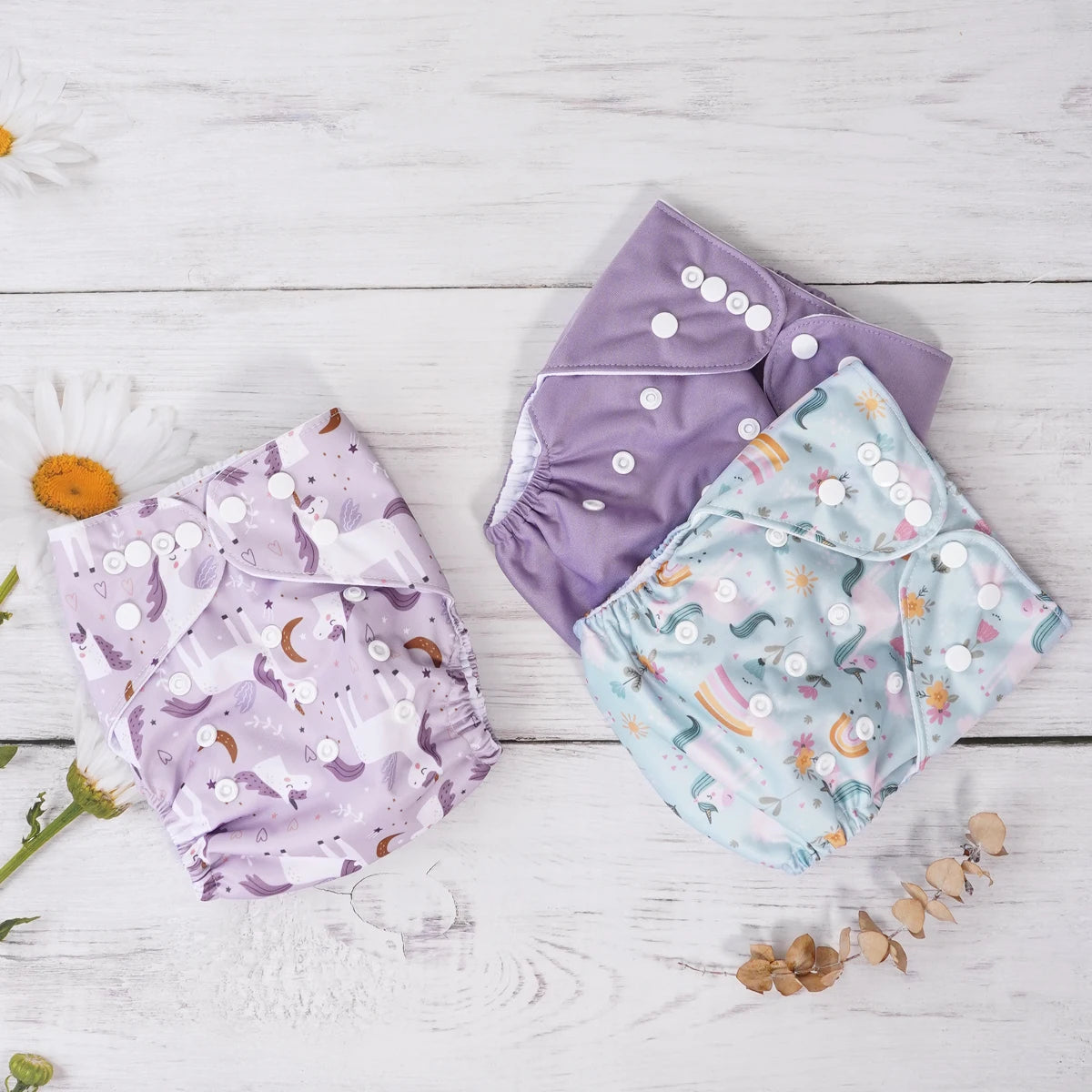 Three reusable cloth baby diapers featuring unicorn, floral, and purple prints with adjustable snap closures.