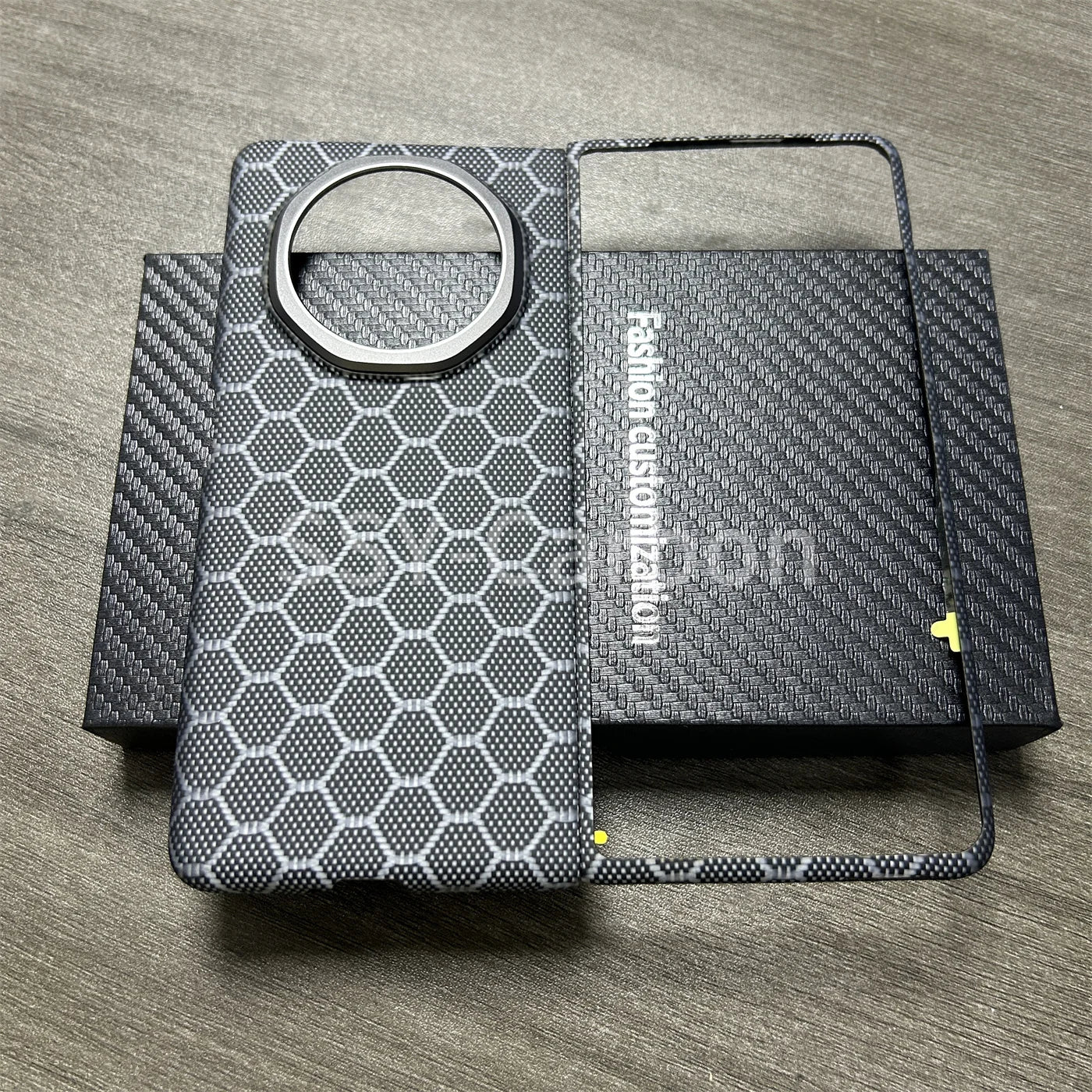 Gray honeycomb-patterned phone case with a circular camera cutout and matching slim frame on a carbon fiber box.