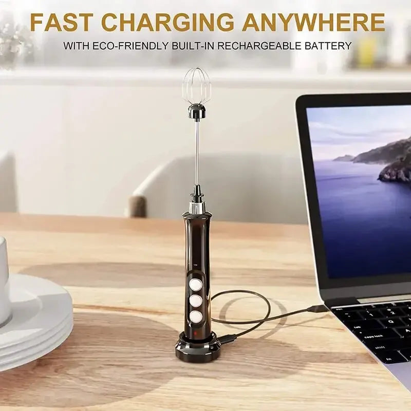 Black rechargeable electric milk frother with metal whisk, charging on a stand next to a laptop.