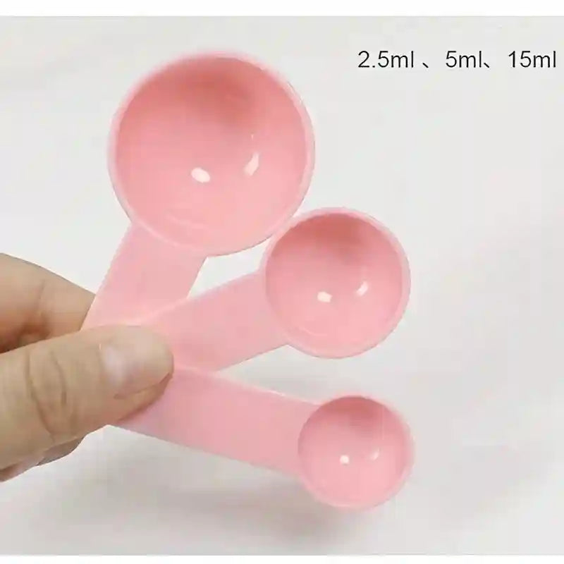 Hand holding three light pink plastic measuring spoons in sizes 2.5ml, 5ml, and 15ml for face mask mixing.
