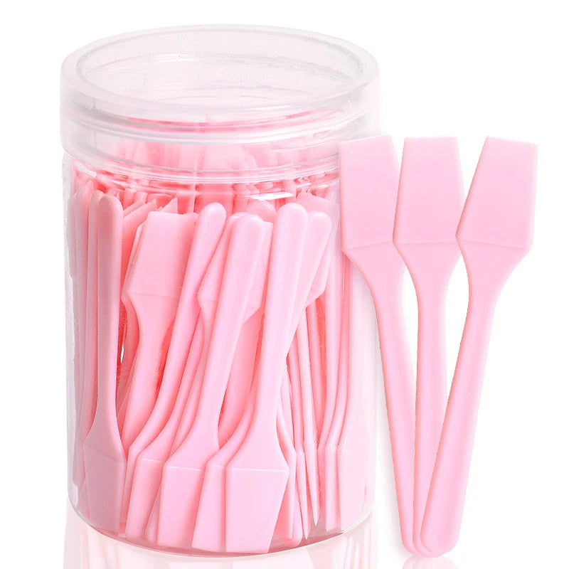 A clear plastic jar filled with 100 small, pink plastic cosmetic spatulas for applying creams and makeup.