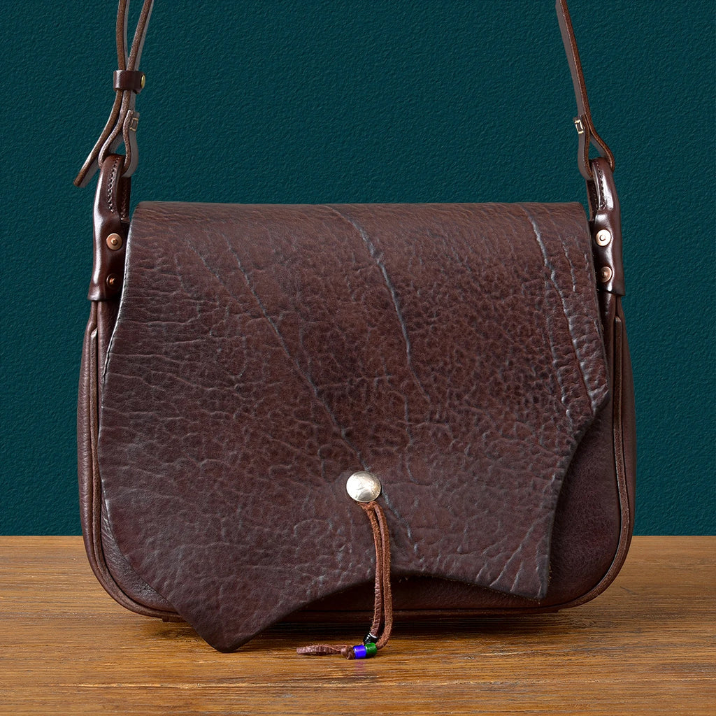 PNDME handmade dark brown genuine cowhide messenger bag with textured flap, asymmetrical cut, and rivet accents.