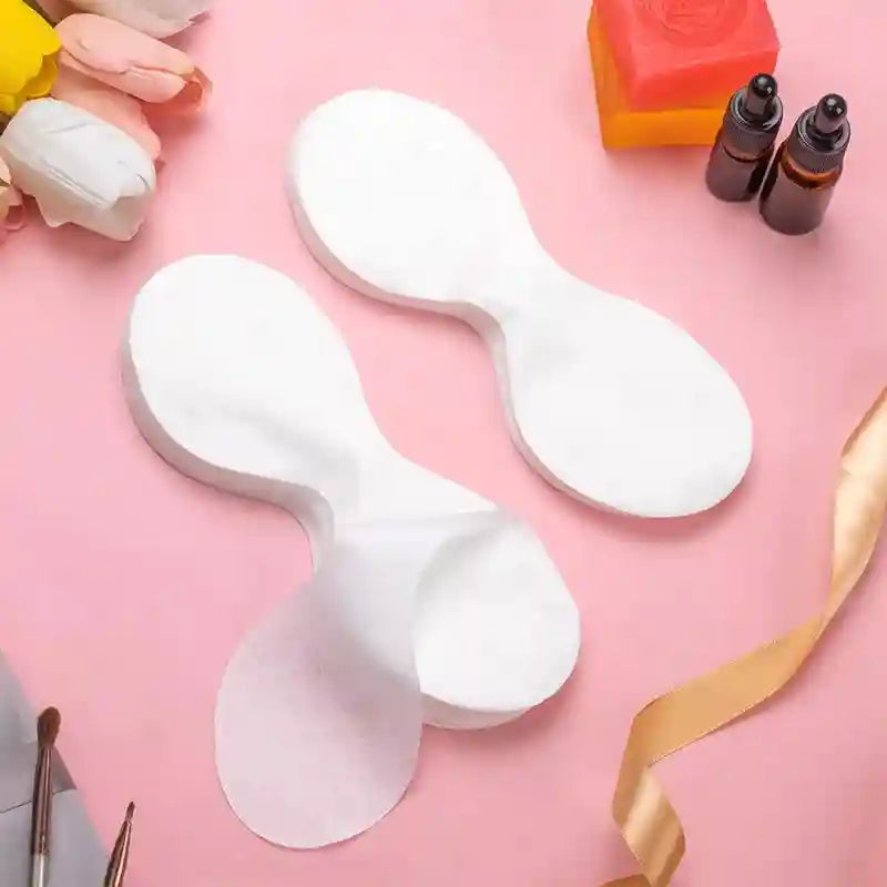 Thin white, disposable cotton eye mask papers shaped like connected ovals, arranged on a pink surface with accessories.