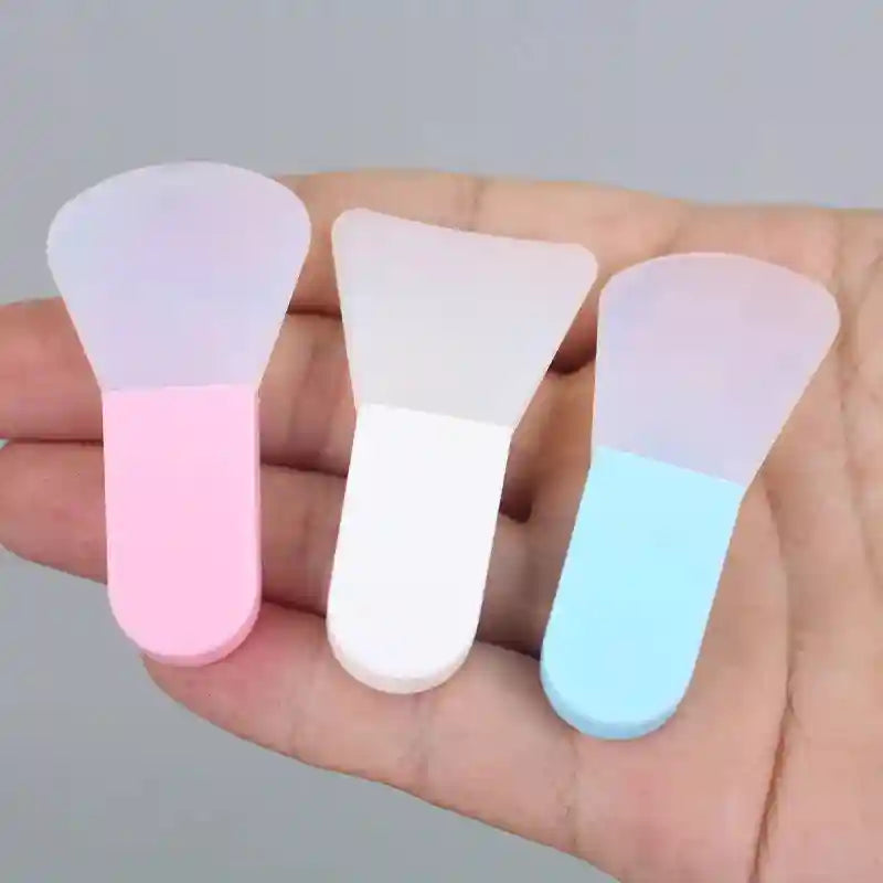 Three silicone face mask spatulas with flexible, translucent heads and pink, white, and blue handles held in a hand.
