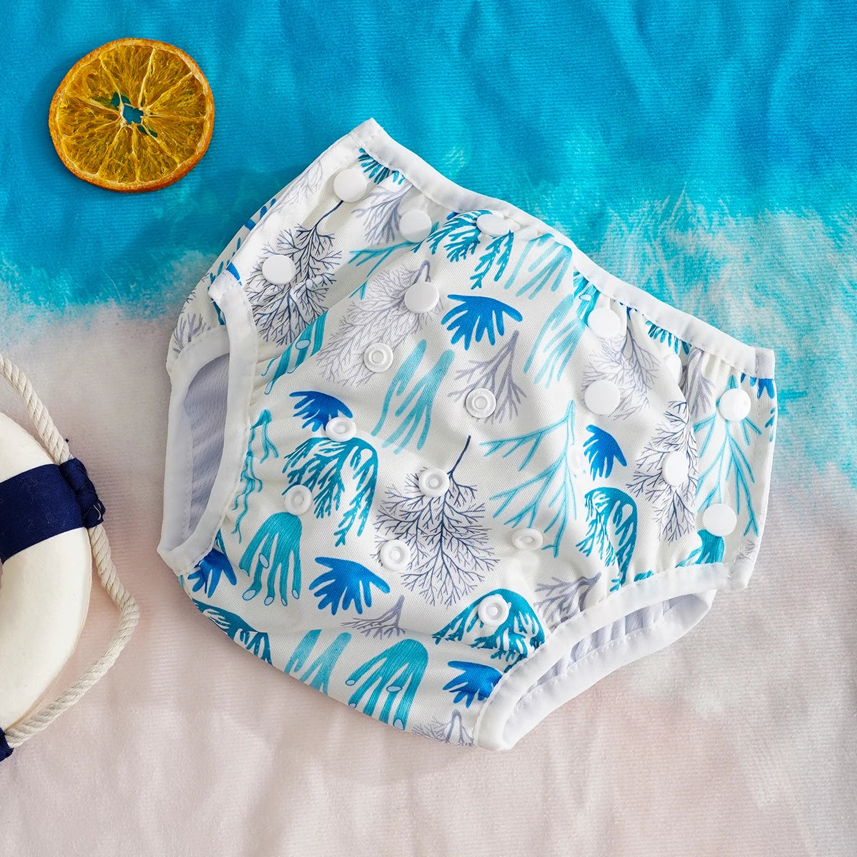 Reusable cloth baby diaper with adjustable snaps and blue coral print design on a beach-themed background.