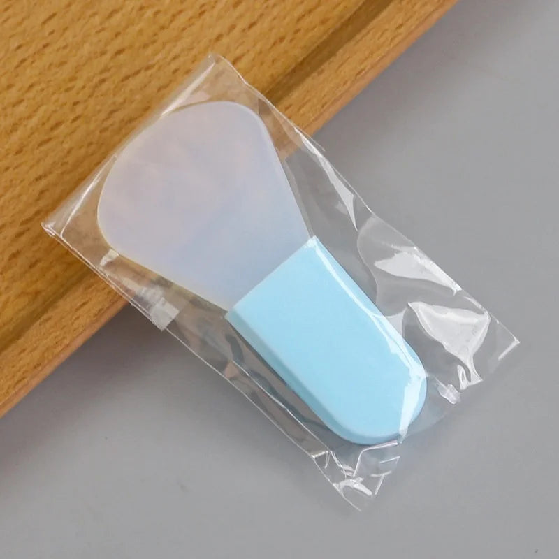 A small, flexible silicone facial mask applicator with a light blue handle in clear plastic packaging.