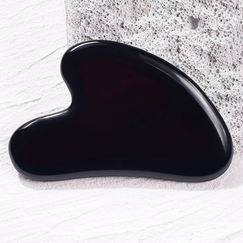 Polished, heart-shaped black stone gua sha facial massage tool resting against a textured gray rock.