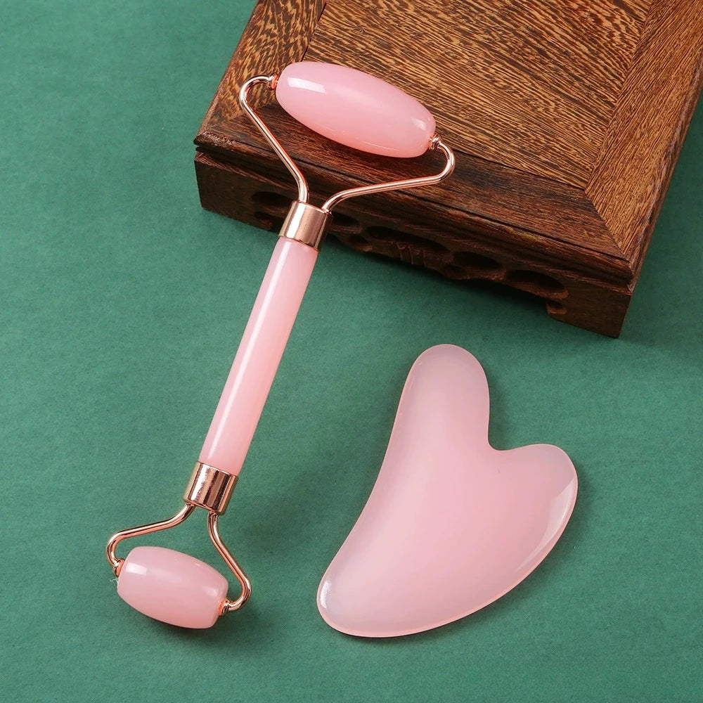 Pink quartz facial roller and heart-shaped gua sha board with rose gold metal accents on a green surface.