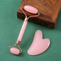 Pink quartz facial roller and heart-shaped gua sha board with rose gold metal accents on a green surface.