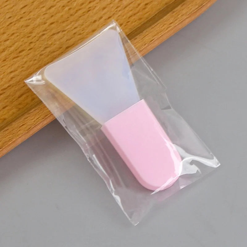 A translucent silicone facial mask applicator with a pink handle, packaged in a clear plastic sleeve.