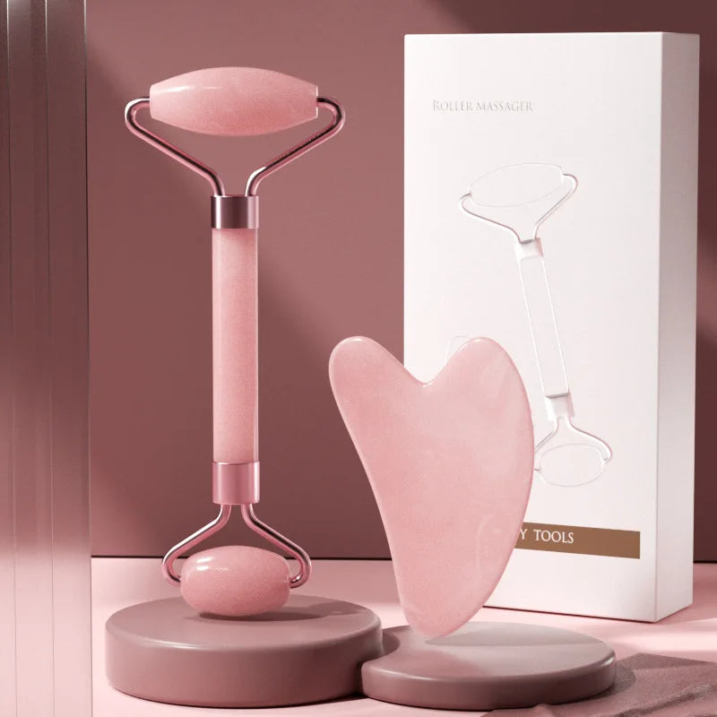 Pink resin facial massage roller and heart-shaped Gua Sha tool set with their white packaging box.