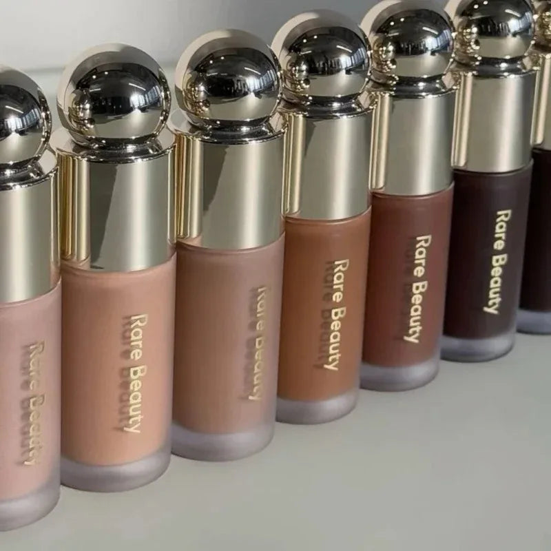 Rare Beauty liquid contour and highlighter bottles in a gradient of nude-to-deep skin tones with metallic gold caps.