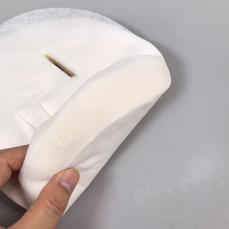 A hand holds an ultra-thin, white, disposable cotton facial mask sheet with pre-cut eye openings.