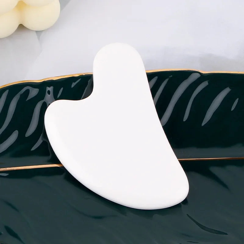 White heart-shaped resin gua sha massage tool resting on a dark green leaf-shaped decorative tray.