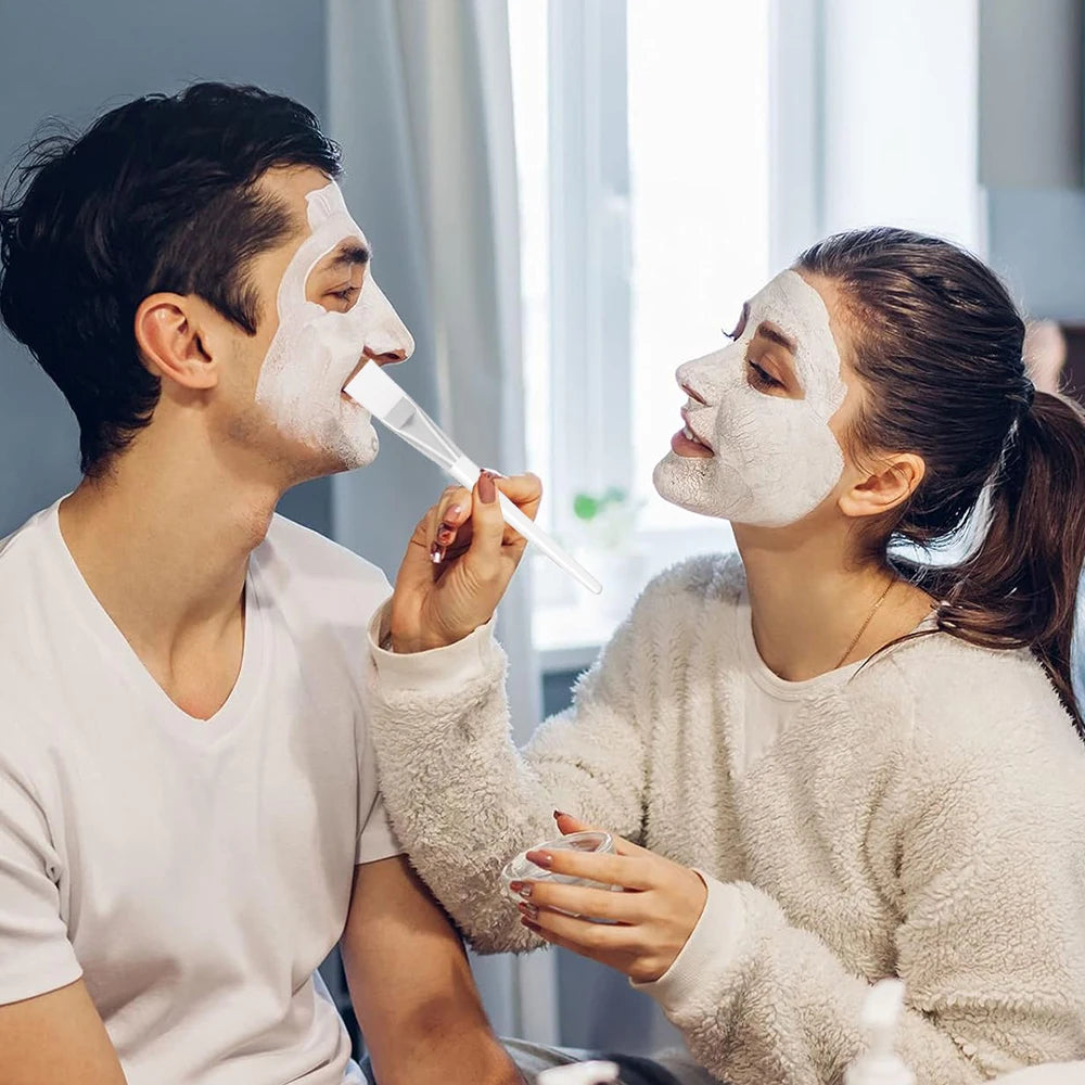 A woman uses a flat, transparent-handled silicone brush to apply a white facial mask onto a man's face.