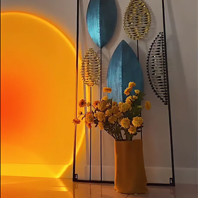 Metal wall art with blue and gold leaf shapes and a yellow vase with flowers in front, lit by a sunset projection.