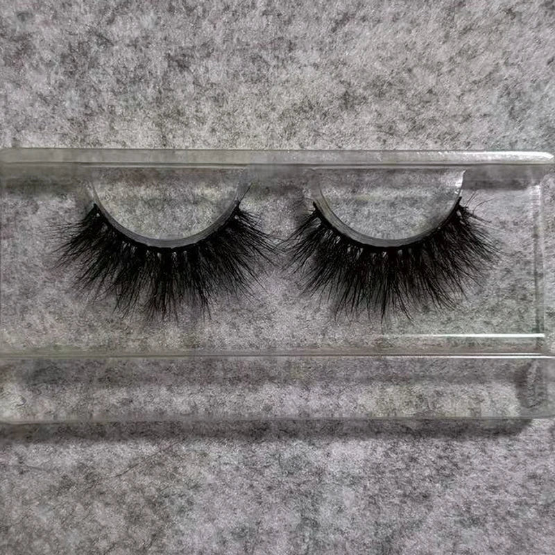 Pair of black 3D mink false eyelashes with a flexible band, packaged in a clear plastic tray on a gray surface.